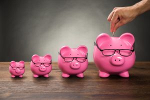 Close-up Of Person Hand Inserting Coin In Pink Piggybank On Table